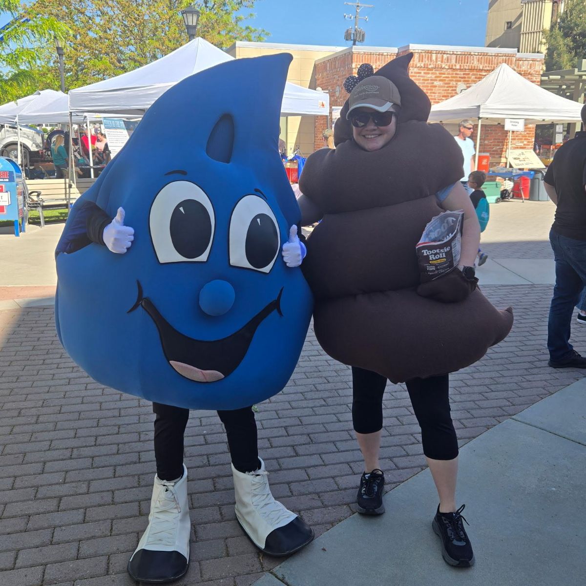 Meridian Public Works' mascots, Hydro (a person dressed up in as a water drop with big eyes, a nose, and smiling face) and Lady Poo (a person dressed up at the poo emoji) posing for a picture in front of event tents